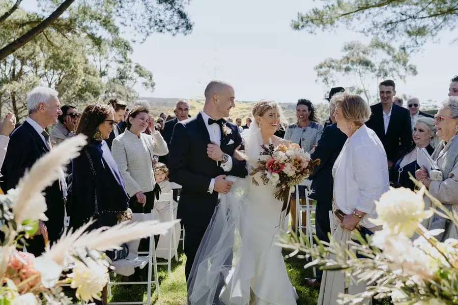 Newlyweds stand among smiling guests beside the outdoor ceremony chairs.