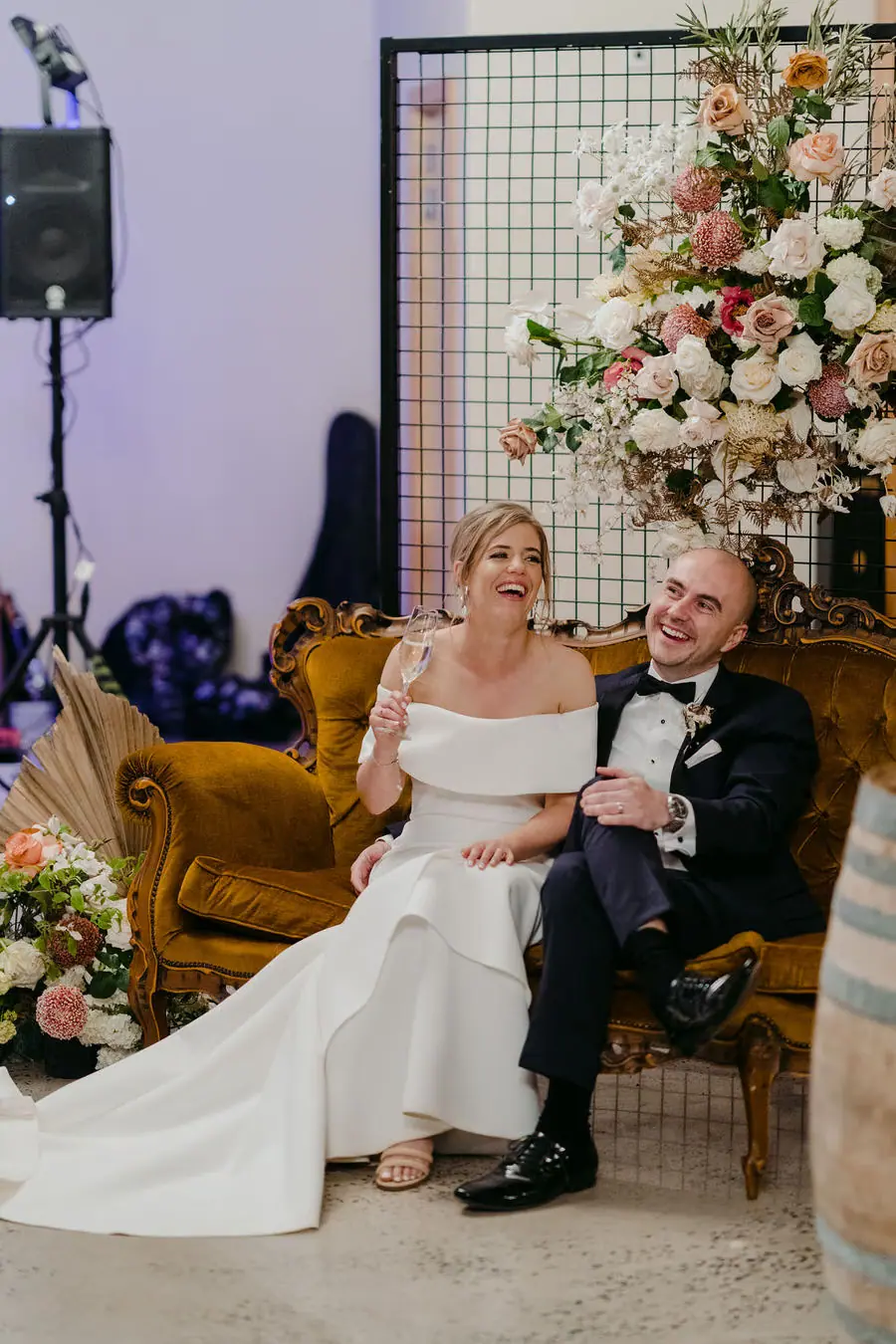 Couple sits together on ornate chairs beside a tall floral arrangement.