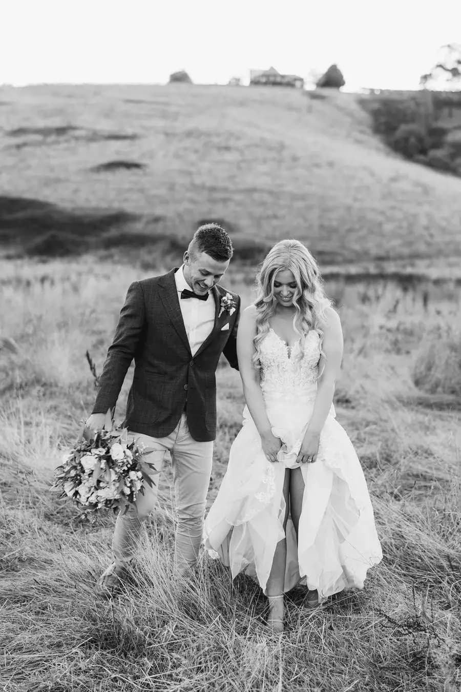 Black and white photograph of the couple walking through a field with the bride gathering her dress.