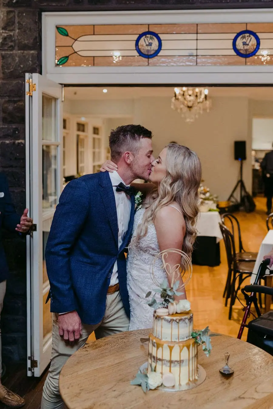 Couple kisses beside a tower of champagne glasses on a wooden table.