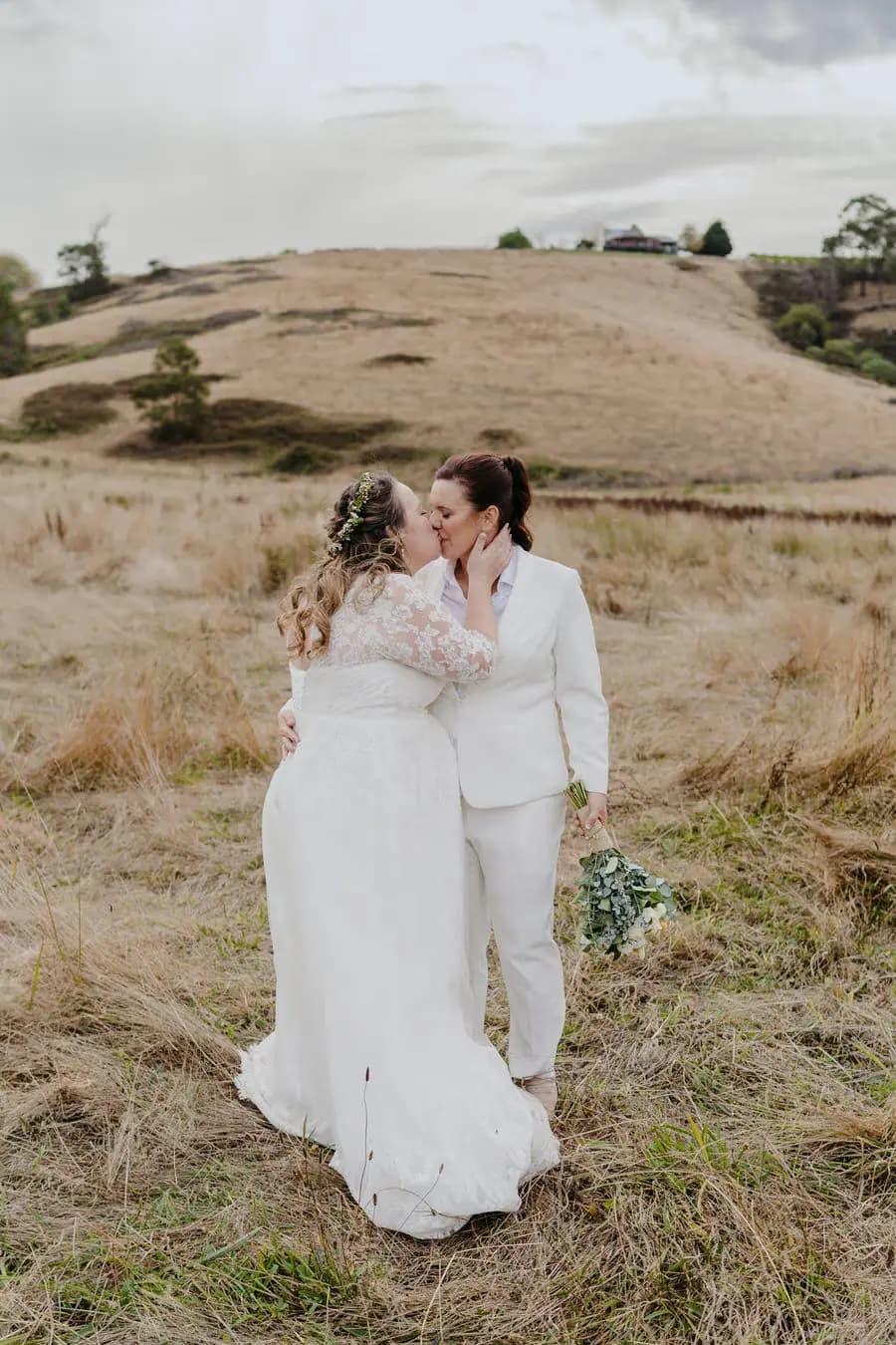 Two brides embrace in a dry field beneath cloudy skies.