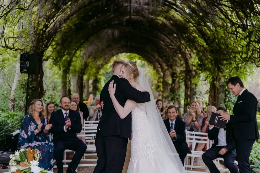 Couple kisses beneath an archway of green vines as guests watch.