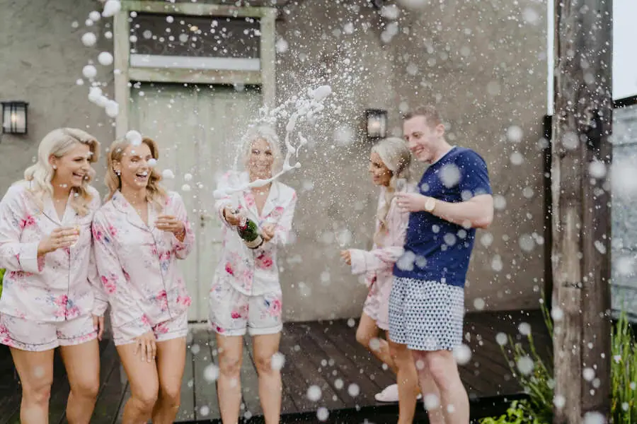 Women laugh as champagne sprays outdoors beside a building.