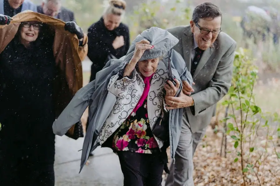 Guests help an older woman shelter beneath a jacket at a rainy wedding.