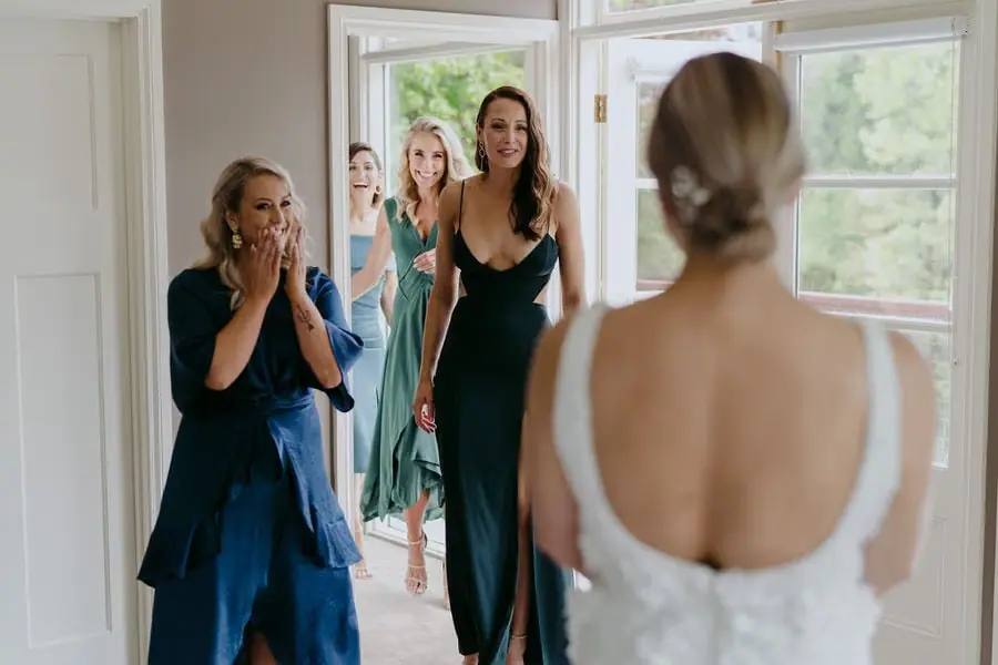 Bridesmaids react to the bride's dress in a bright room.