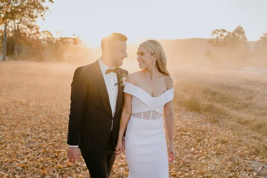 Couple walks hand in hand through a sunlit dry field.