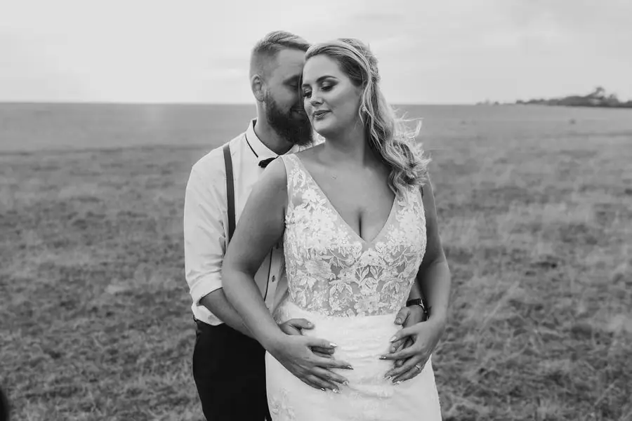 Black and white photograph of the groom embracing the bride from behind in a field.