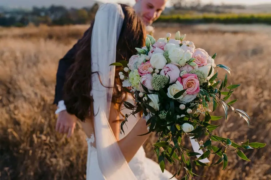 Couple kisses behind a large pink and white bouquet in a field.