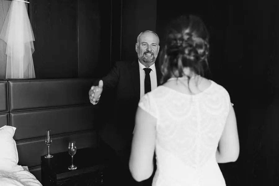 Black and white photograph of an older man opening his arms toward the bride indoors.