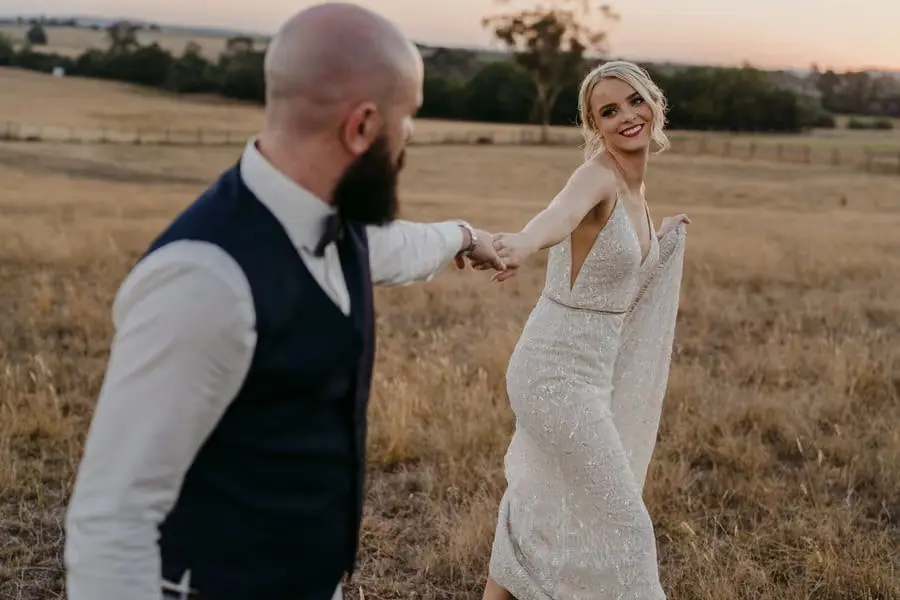 Groom leads the smiling bride by the hand across a dry field.