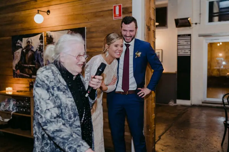 Older guest holds a microphone while the couple laughs beside her.