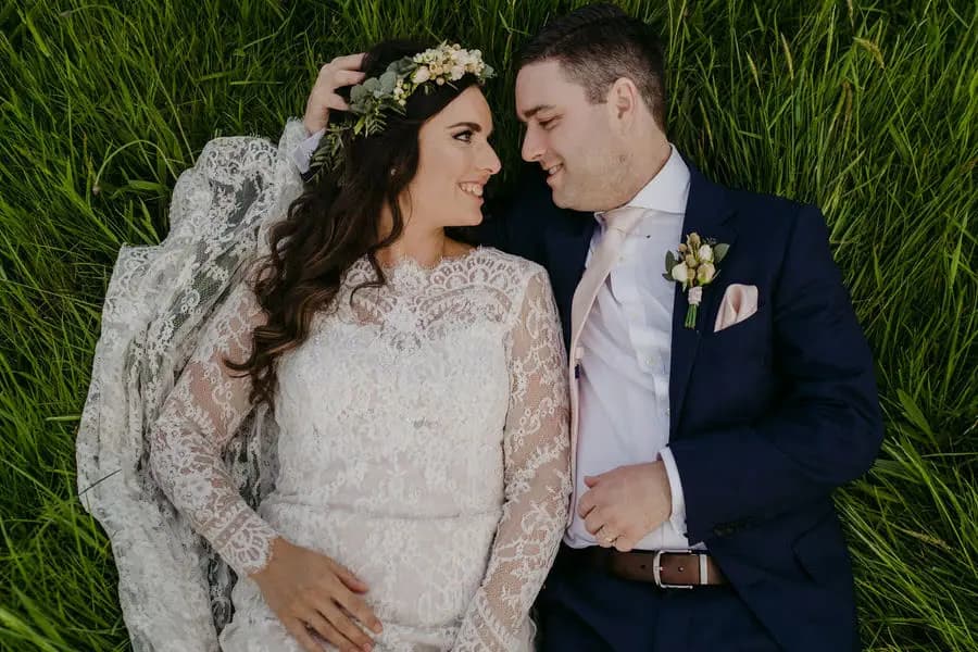 Couple lies side by side in green grass smiling toward each other.