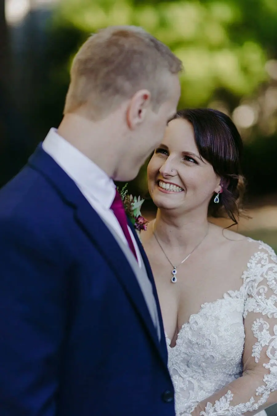 Bride smiles up toward the groom beneath garden trees.