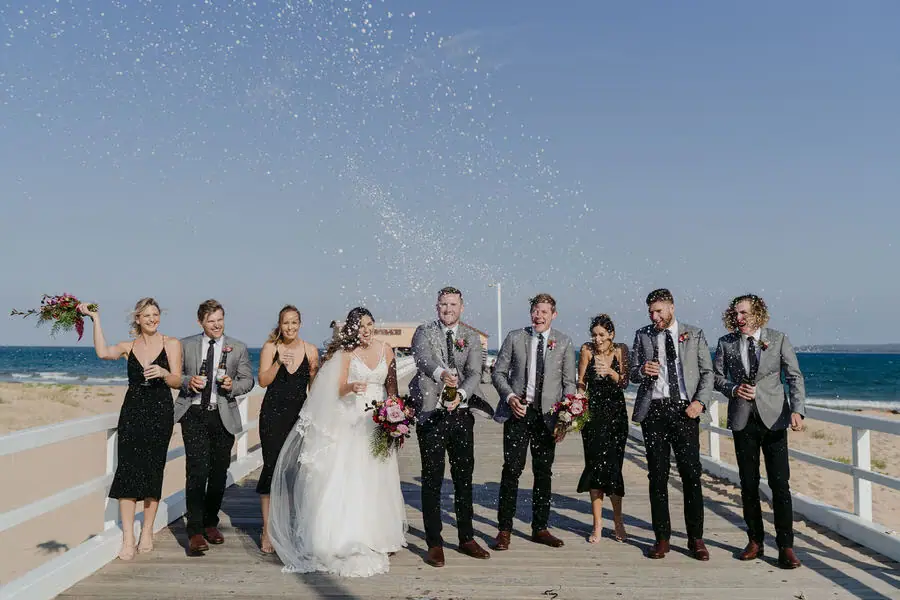 Wedding party celebrates beside the ocean beneath airborne champagne spray.