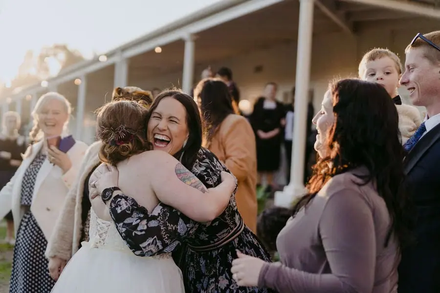 Bride embraces a laughing guest outside the wedding venue.