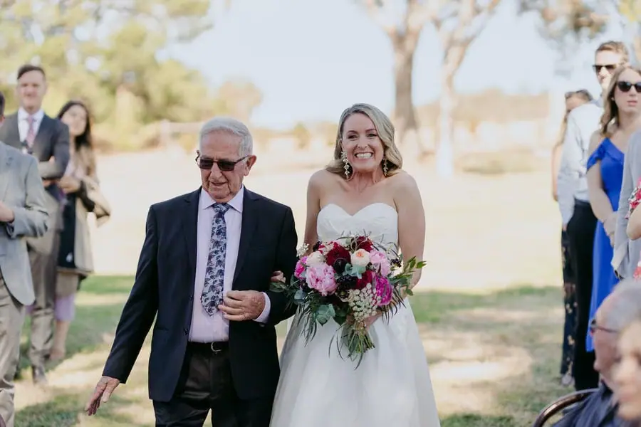 Bride walks arm in arm with an older man through outdoor wedding guests.