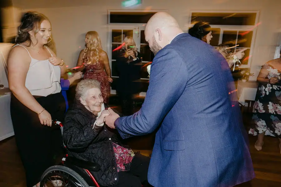 Groom holds the hands of an older guest in a wheelchair on the reception dance floor.