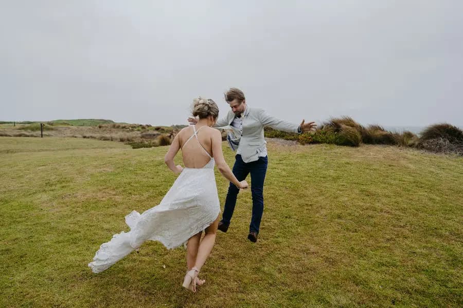 Couple dances playfully on a grassy coastal hill with the bride's dress blowing sideways.