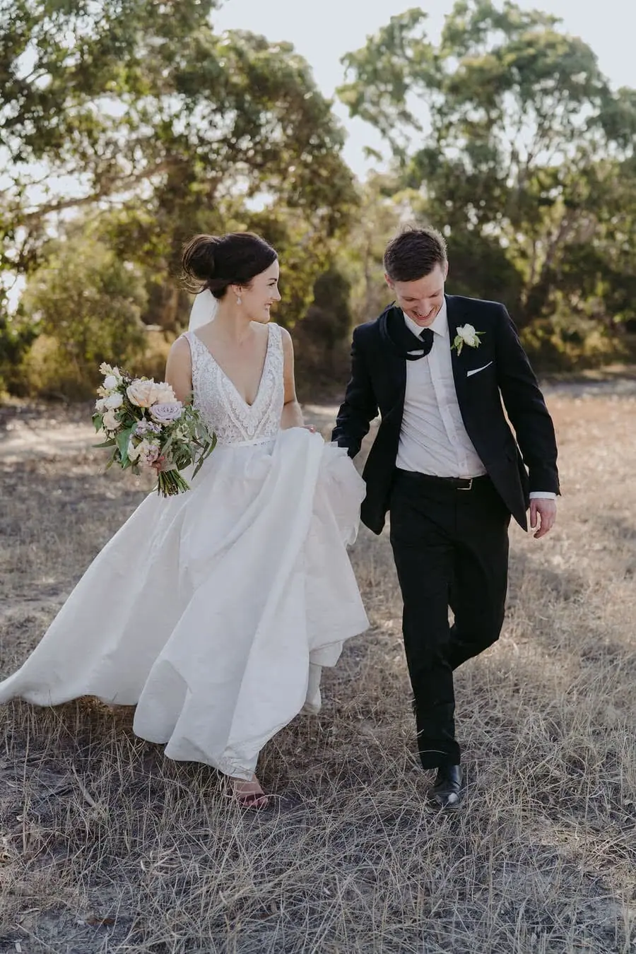 Couple walks through dry grass with the bride lifting her skirt.