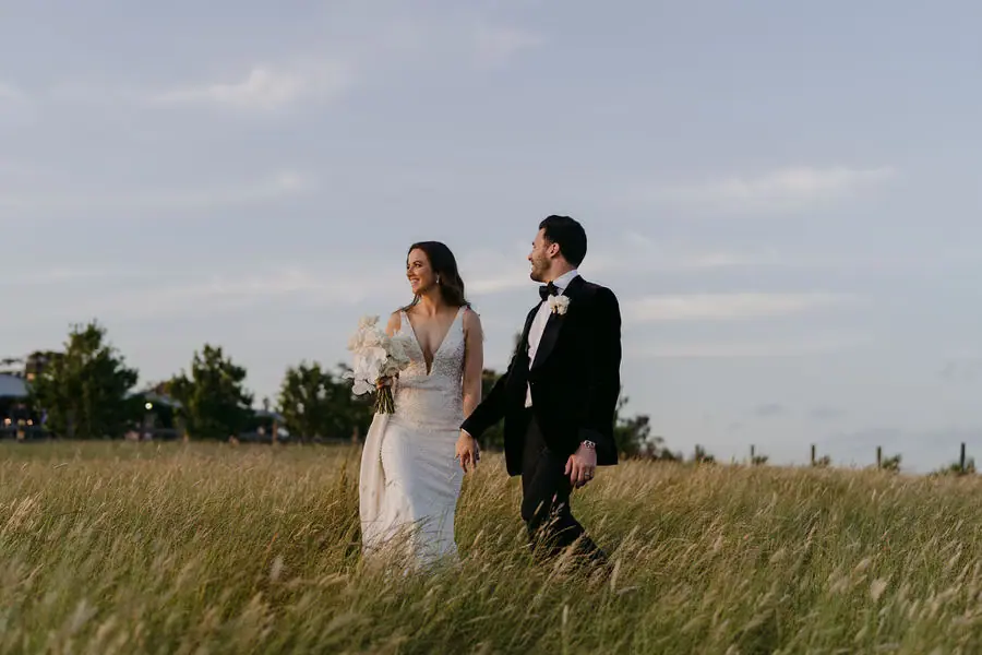 Couple walks hand in hand through tall grass beneath an evening sky.