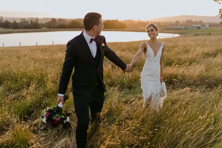 Groom leads the smiling bride by the hand through a sunlit field beside water.