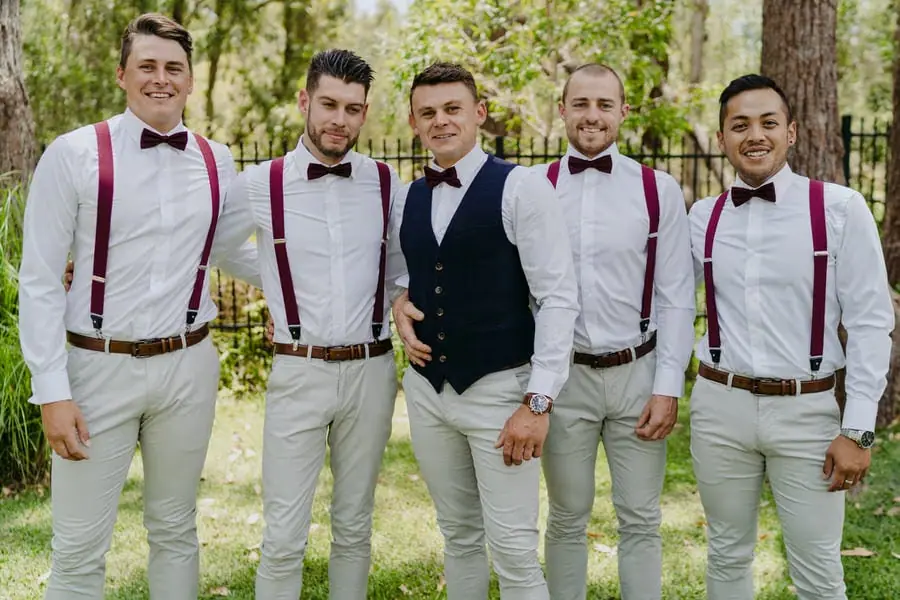 Groom and four groomsmen posing together beneath trees in bow ties and burgundy braces
