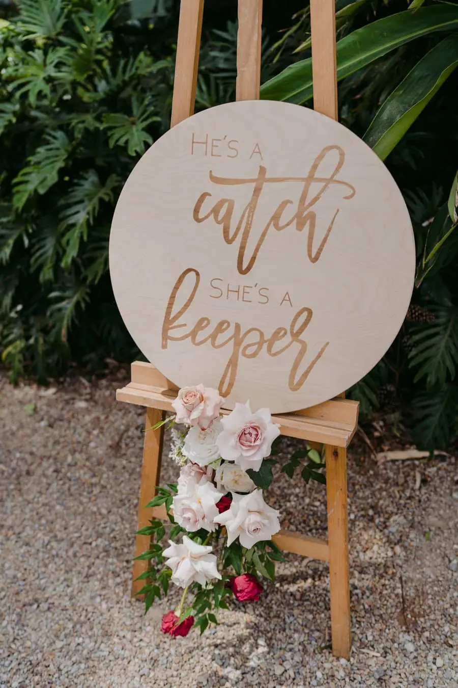 Round wooden wedding sign decorated with pale flowers beside a garden path