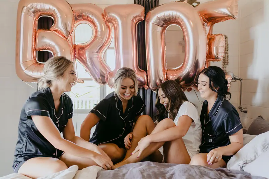 Bride and bridesmaids laughing on a bed beneath large BRIDE balloons