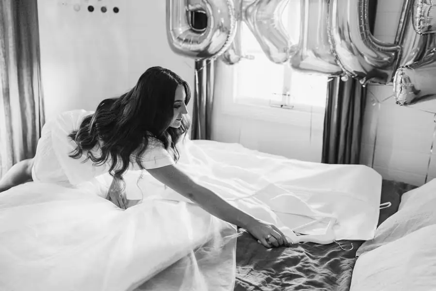 Black and white photograph of the bride reaching for white shoes beside her dress on a bed