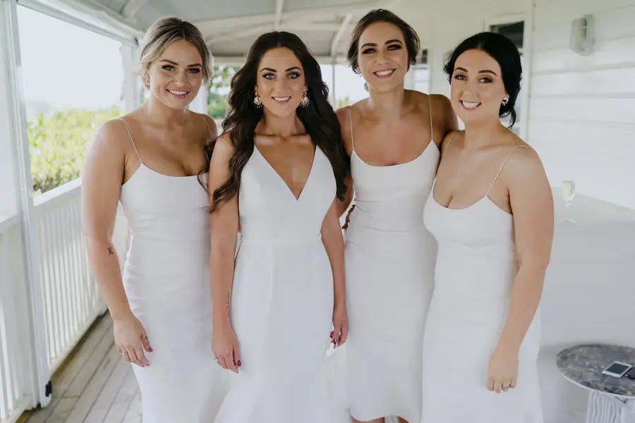 Bride and bridesmaids smiling together in white dresses on a veranda