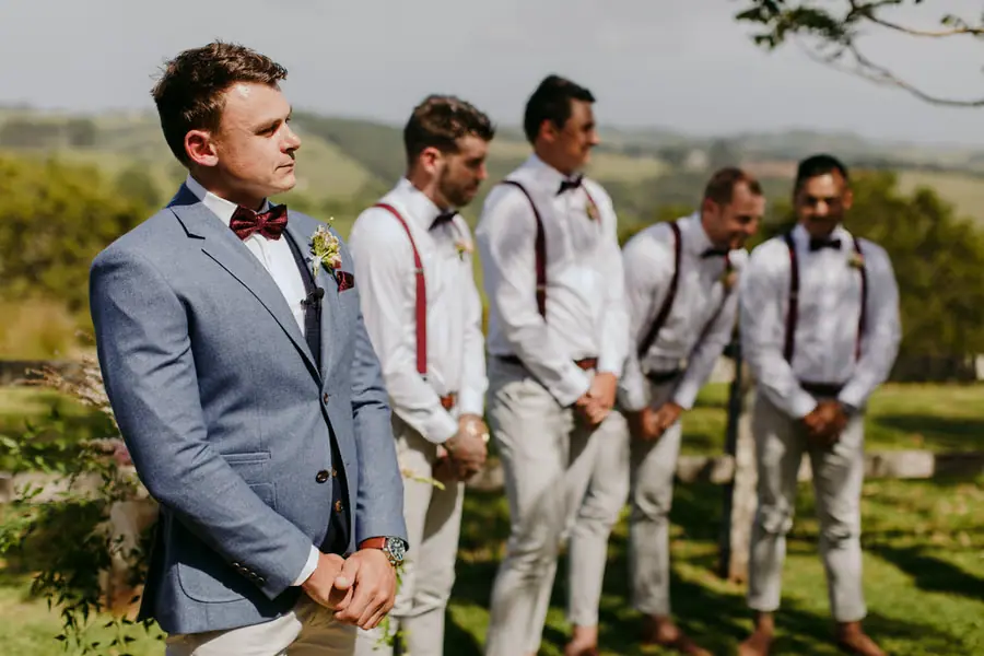 Groom and groomsmen waiting beside the ceremony flowers with green hills behind them