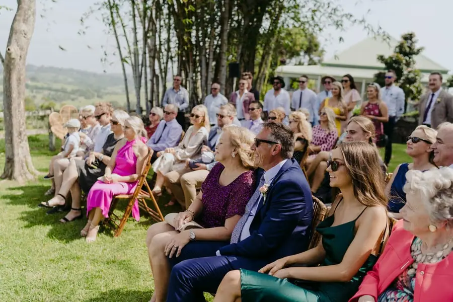 Guests seated in rows of wooden chairs beneath trees at the ceremony