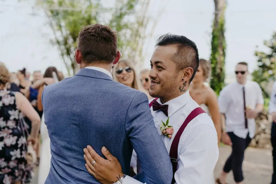 Groom greeting a groomsman wearing burgundy braces outdoors