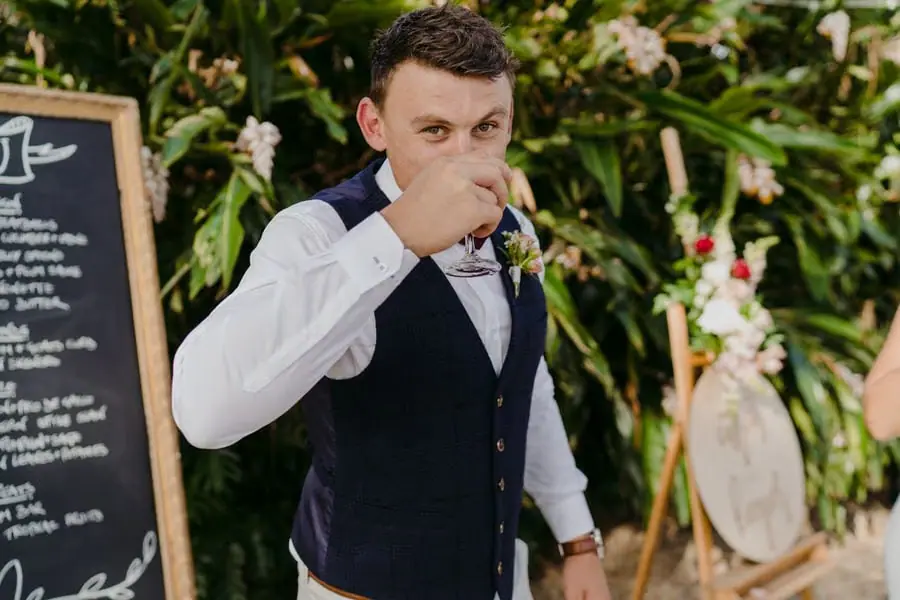 Groom drinking from a glass beside floral decorations and a menu board