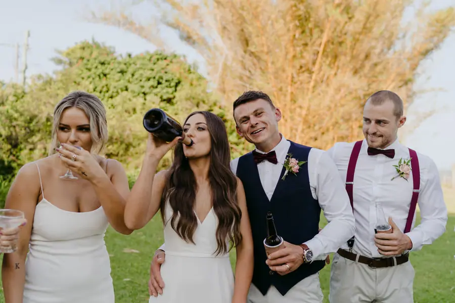 Bride drinking from a bottle while wedding party members laugh beside her