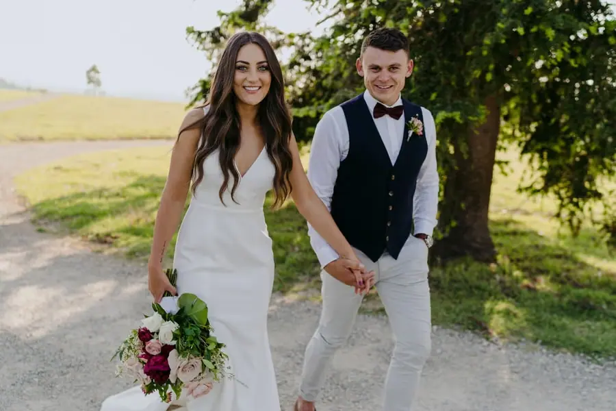 Bride and groom smiling toward the camera while holding hands beside a country lane