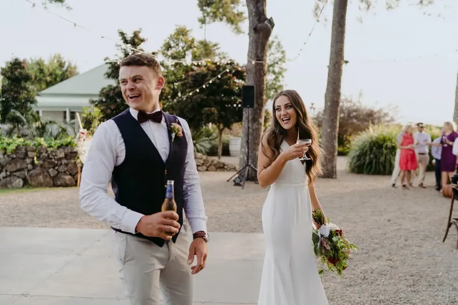 Bride and groom smiling with drinks beside a stone wall and reception garden
