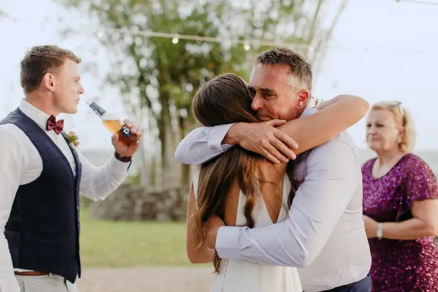 Bride hugging a man in a white shirt while the groom holds a drink beside them
