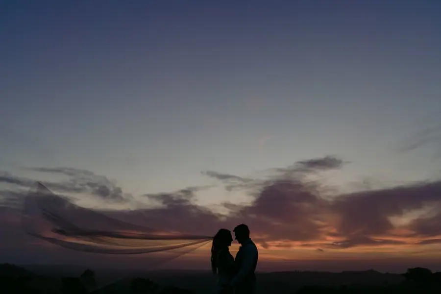 Silhouettes of the bride and groom leaning together beneath a colourful sunset sky