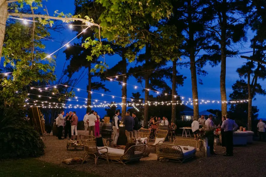 Outdoor wedding reception beneath strings of festoon lights and tall trees at dusk