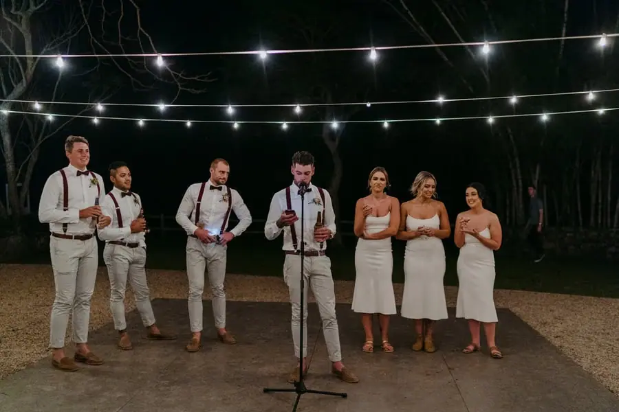Wedding party standing behind a microphone beneath outdoor festoon lights