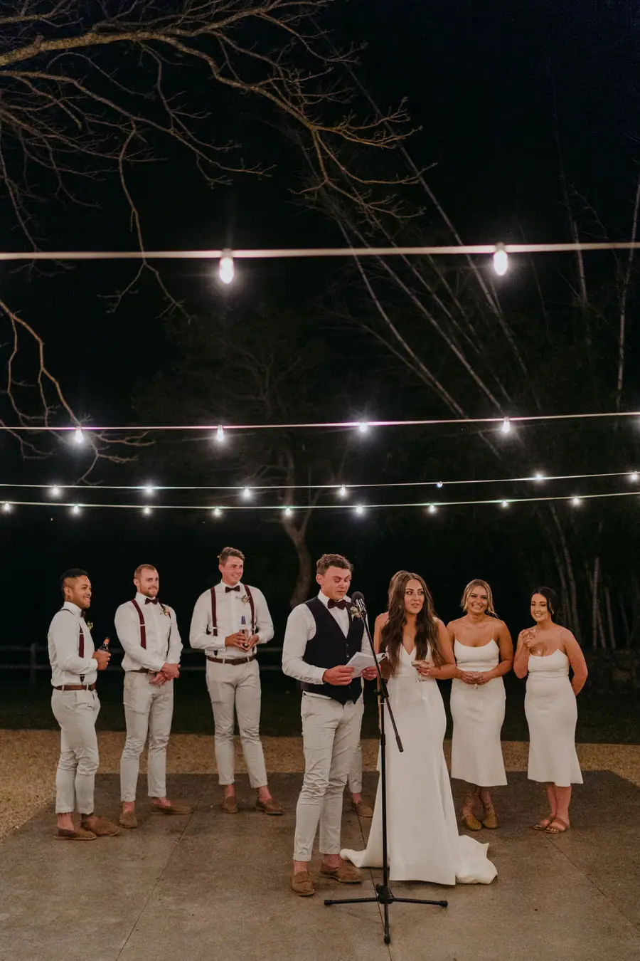 Bride and groom speaking beside a microphone with the wedding party behind them