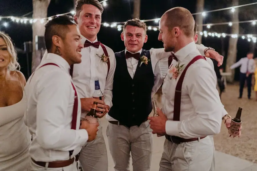 Groom and groomsmen laughing together with drinks beneath festoon lights