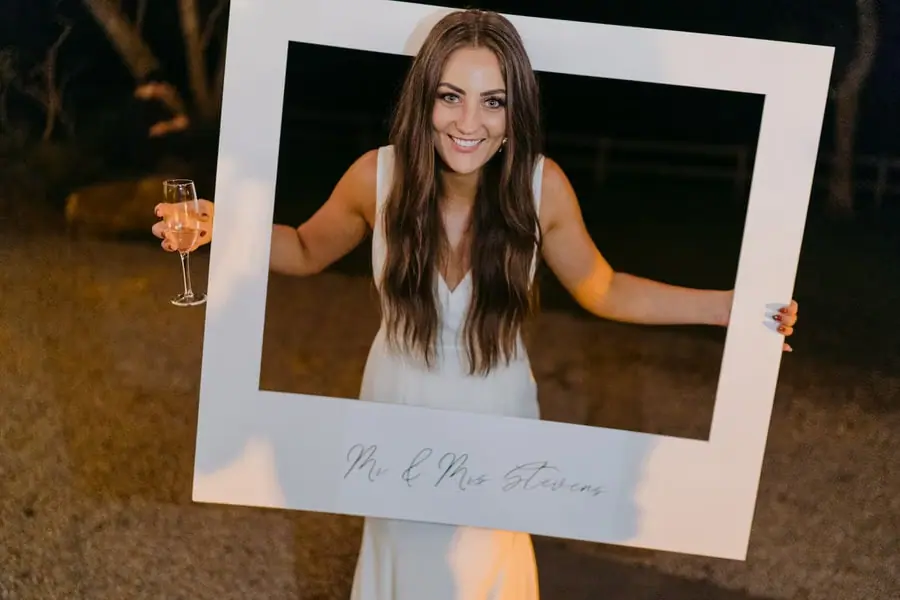 Bride smiling through a large white photo frame while holding a glass