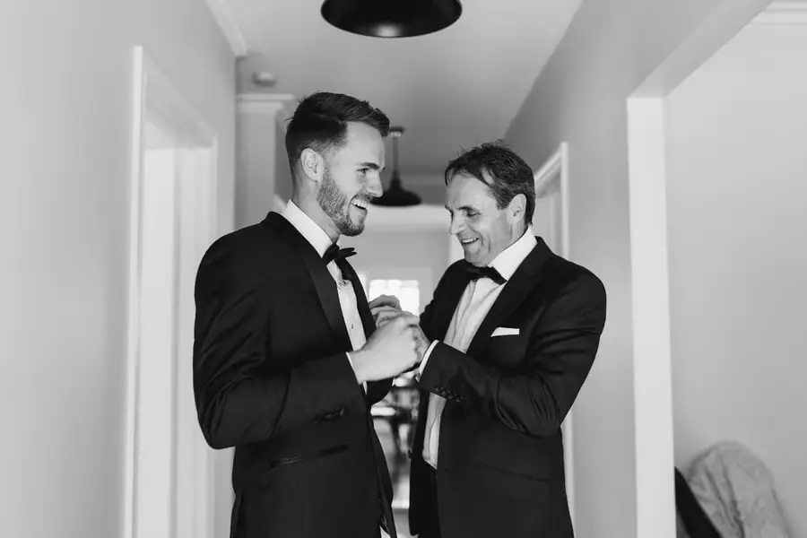 Black-and-white photograph of two men laughing while adjusting their suit jackets