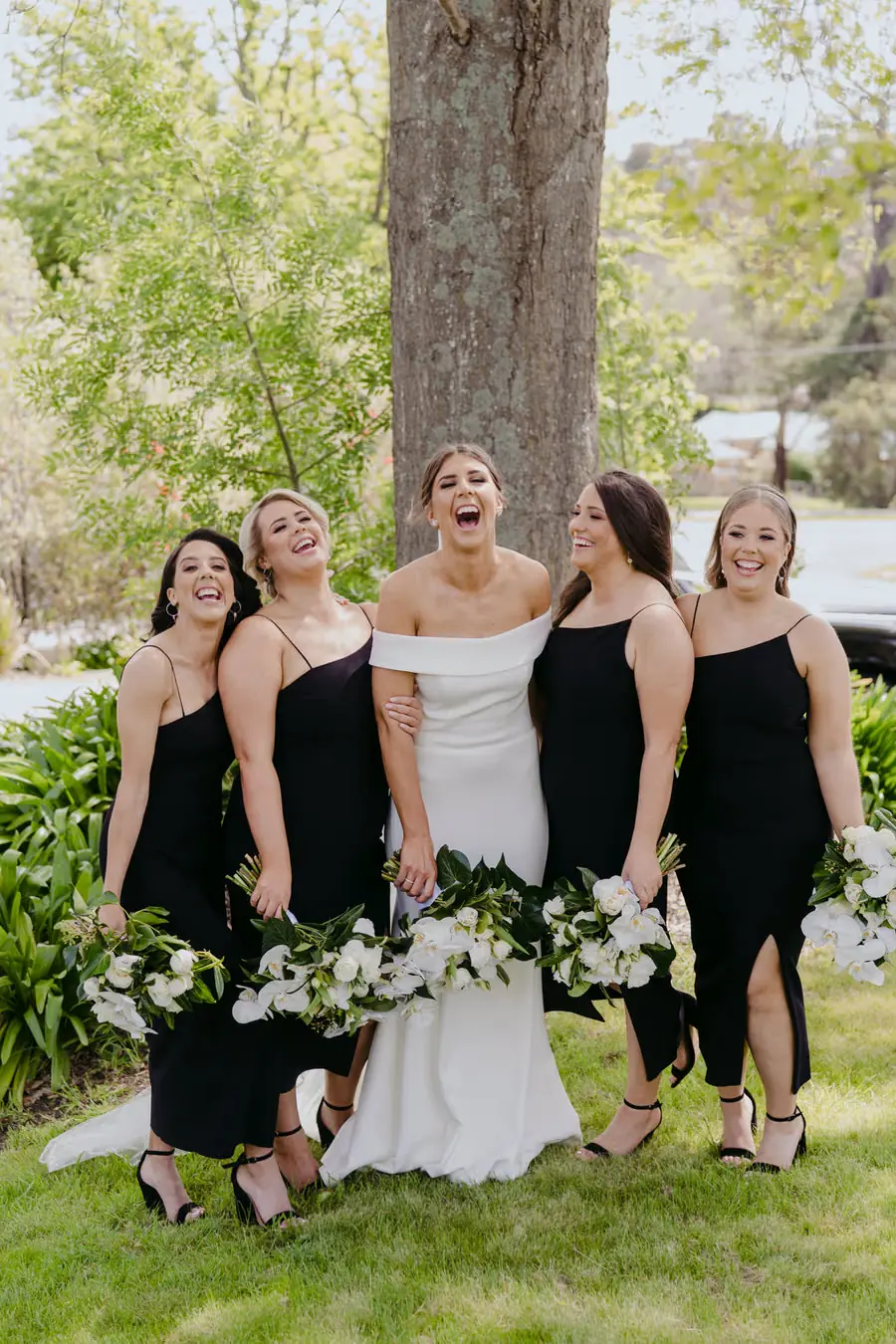 Bride and four bridesmaids laughing together with white bouquets beneath a tree