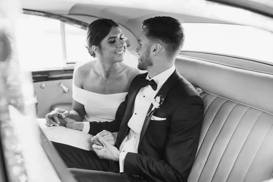 Black-and-white photograph of the bride and groom holding hands in the back seat of a car