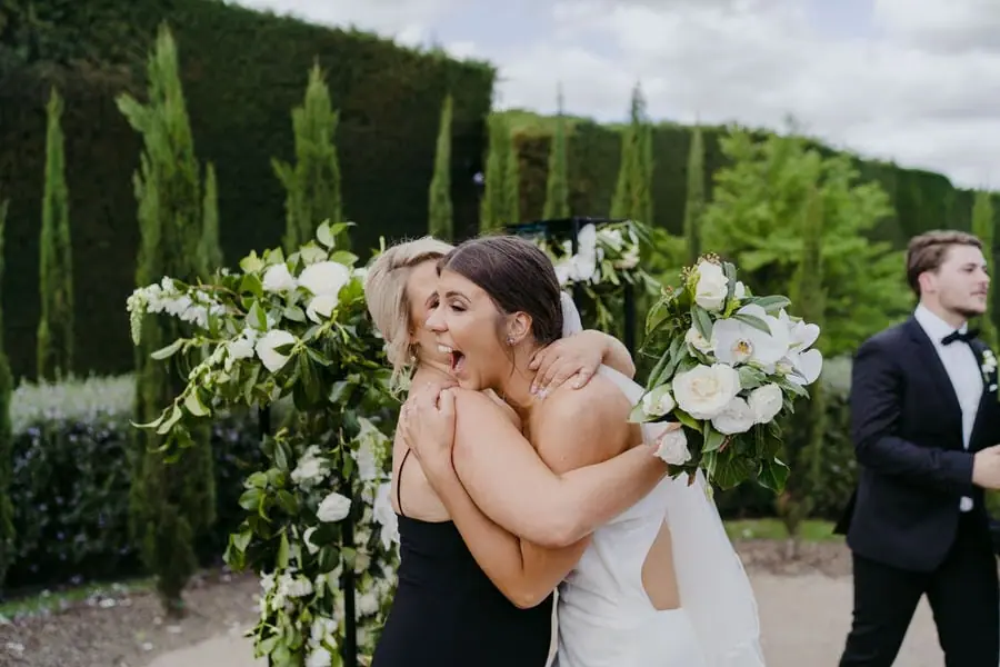 Bride hugging a woman beside a white floral arrangement