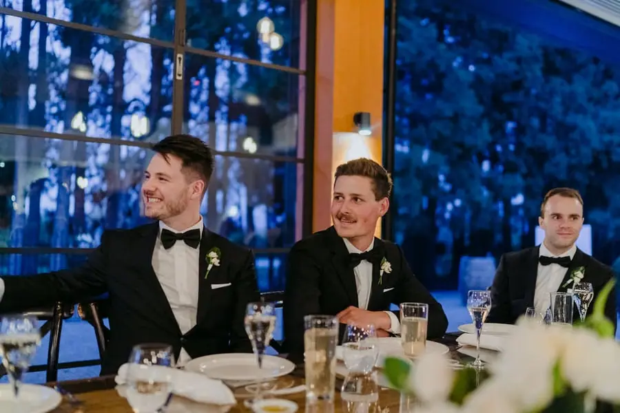 Three men smiling together at a reception table beside large windows