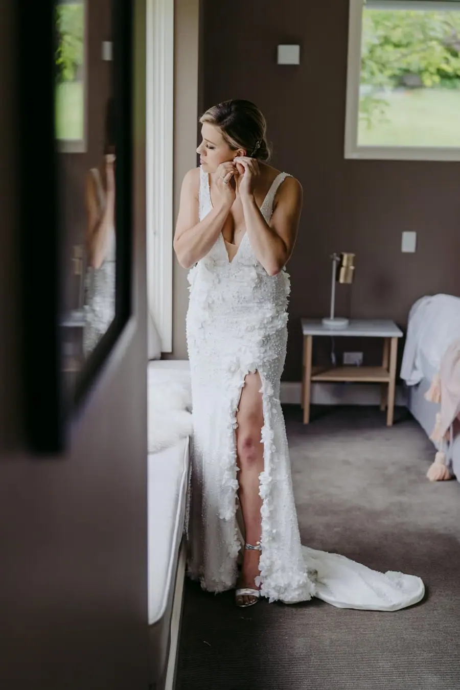 Bride adjusting an earring beside a bedroom mirror.